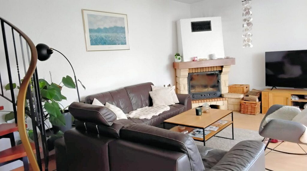 Auf dem Foto ist ein modernes Wohnzimmer zu sehen. In der hinteren Ecke befindet sich ein offener Kamin. Dieser ist im unteren Bereich mit großen hellen Steinen und Klinkern eingefasst, im oberen Bereich des Kamins befindet sich ein Lüftungsgitter. Rechts neben dem Kamin ist ein Sitzplatz integriert, darauf steht ein geflochtener Korb und daneben -allerdings auf dem Fußboden- ein ebenfalls geflochtener Korb mit Holzscheiden. Im Anschluss erkennt man ein TV Schrank mit TV-Gerät. Links neben dem Kamin befindet sich eine dunkelbraune Leder-Sitzecke mit hellen Kissen und einem hellen Feld auf der Sitzfläche. Vor der Sitzgruppe steht ein Holztisch mit filigranen Metallbeinen und Ablage. Der Fußboden besteht aus hellem Laminat und unter der Sitzgruppe ist ein grauer Teppich. Des Weiteren erkennt man gegenüber der Sitzgruppe ein Teil eines Schaukelsessels. Im linken vorderen Bildrand ist ein Teil einer Wendeltreppe zu sehen, sowie eine Pflanze und eine Leselampe.