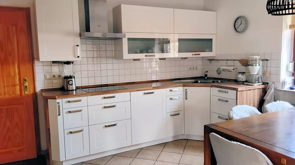 Das Bild zeigt eine helle und moderne Küche. Der Hauptfokus liegt auf den weißen Küchenschränken, die in einer L-Form angeordnet sind. Die Oberschränke haben teilweise Glastüren, durch die man einige Gegenstände erkennen kann. Die Arbeitsplatte besteht aus hellem Holz, was einen schönen Kontrast zu den weißen Fronten bildet. Linkerhand im Bild ist eine hölzerne Tür mit goldenen Beschlägen zu sehen. Darauf folgend im Arbeitsbereich steht ein silberner Standmixer auf der Arbeitsplatte. Die Wand hinter der Arbeitsplatte ist mit weißen, rechteckigen Fliesen verkleidet, die ein Schachbrettmuster ergeben. Eine Edelstahlabzugshaube ist über dem Kochfeld angebracht. Rechts im Bild schließt sich die Küchenzeile an. Dort befinden sich ein Spülbecken, Armatur und ein weißer Kaffeemaschine auf der Arbeitsplatte. Eine Wanduhr mit silbernem Rahmen und schwarzen Ziffern hängt an der weißen Wand über dem Spülbereich. Daneben sind einige Küchenutensilien an einer Stange aufgehängt, darunter ein Papierrollenhalter und ein Geschirrtuch. Im Vordergrund ist ein Teil eines großen Holztisches zu sehen, der mit zwei Stühlen belegt ist. Die Stühle sind mit hellen, flauschigen Bezügen versehen. Der Tisch und die Stühle sind in einem warmen Holzton gehalten. Die Beleuchtung im Raum ist hell und natürlich, was durch ein Fenster rechts im Bild angedeutet wird. Die Komposition ist so gewählt, dass sie einen guten Überblick über die Küchenzeile und den Essbereich gibt. Die Aufnahme ist leicht von oben und schräg gemacht.