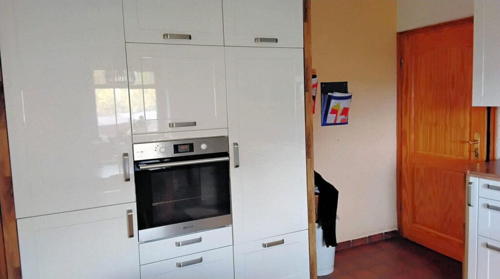Das Bild zeigt eine Küche mit modernen weißen Hochglanzschränken, die an der Wand angebracht sind. In den Schrank integriert ist ein Einbauofen aus Edelstahl mit einem digitalen Display, das die Uhrzeit 12:39 anzeigt. Über dem Ofen befinden sich weitere Schranktüren mit silbernen Griffen. Links neben dem Ofen sind die Schränke hochglänzend weiß, und man kann Reflexionen von Fenstern und möglicherweise der Außenwelt erkennen, was auf eine helle Umgebung hindeutet. Rechts von den weißen Küchenschränken befindet sich eine Holztür aus hellem Holz mit einer deutlichen Maserung und einem goldenen Türgriff. Die Tür ist geschlossen und steht im Kontrast zu den modernen weißen Küchenmöbeln. An der Wand neben der Tür hängt ein blauer Zeitschriftenständer mit verschiedenen farbigen Blättern oder Magazinen. Die Wand selbst ist in einem warmen, hellen Orangeton gestrichen. Vor der Wand und neben den Küchenschränken steht ein weißer Mülleimer, teilweise verdeckt von einem schwarzen Kleidungsstück, das über einen Korb gehängt ist. Der Boden ist mit dunkelroten oder braunen Fliesen ausgelegt.