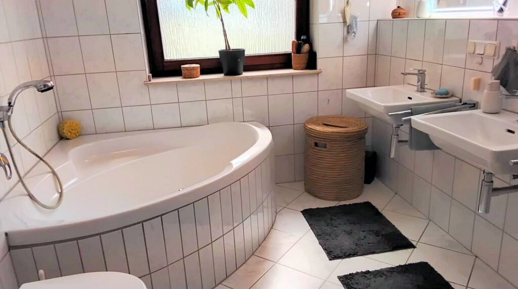 Das Bild zeigt ein Badezimmer. Im Vordergrund links ist eine weiße, eckige Badewanne mit geschwungenen Rändern zu sehen. Sie ist teilweise mit kleinen, hellgrauen Fliesen verkleidet. An der Wand über der Wanne befinden sich größere, quadratische, weiße Fliesen mit grauen Fugen. Ein Duschkopf mit Schlauch ist seitlich an der Wanne angebracht. Links neben der Wanne liegt ein gelber Badeschwamm. Im Hintergrund des Bildes befindet sich ein Waschbeckenbereich mit zwei nebeneinander angeordneten, weißen Waschbecken. Beide Waschbecken sind schlicht und modern gestaltet und haben verchromte Armaturen. Unter den Waschbecken sind silberne Rohre und Handtuchhalter zu erkennen. Zwischen den beiden Waschbecken und links davon steht ein runder Wäschekorb aus hellem, geflochtenem Material mit einem Deckel. Daneben liegen zwei dunkelgraue Teppiche auf dem hell gefliesten Boden. An der Wand über den Waschbecken sind weitere weiße Fliesen angebracht. Rechts oben im Bild ist ein Fenster mit einer Milchglasscheibe und einem dunkelbraunen Rahmen zu sehen. Auf der Fensterbank steht eine Topfpflanze mit grünen Blättern in einem schwarzen Topf, ein kleiner runder Behälter aus geflochtenem Material und ein weiterer Behälter, der Haarbürsten enthält. Auf der Fensterbank rechts befinden sich weitere Dekorationsgegenstände. Die Beleuchtung im Raum ist hell und gleichmäßig, was auf Tageslicht durch das Fenster und/oder künstliche Beleuchtung schließen lässt. Die Farbpalette des Badezimmers ist überwiegend hell, mit viel Weiß und Grau, akzentuiert durch das Braun des Fensterrahmens und des Wäschekorbs sowie das Gelb des Schwamms und das Grün der Pflanze.