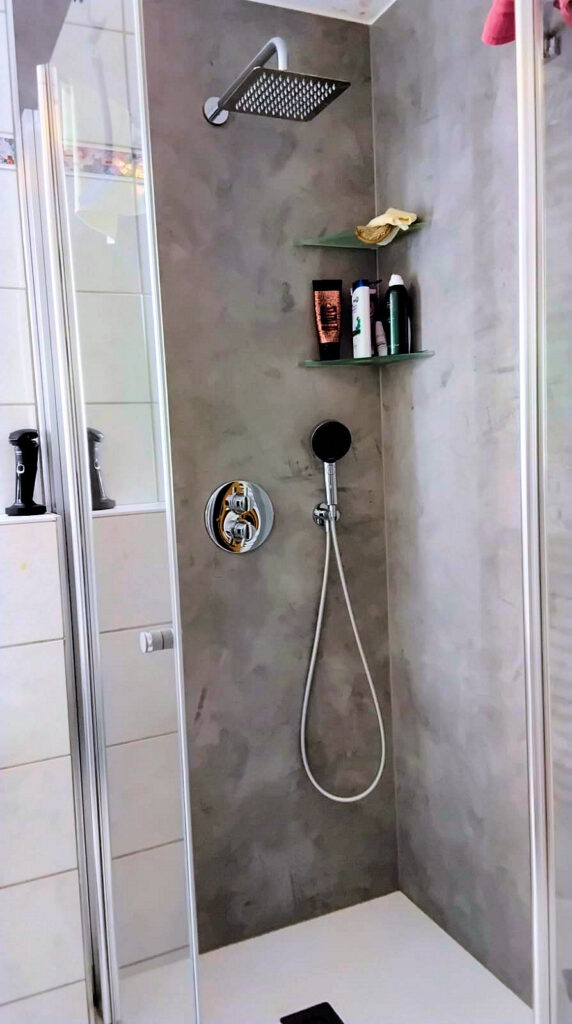 Auf dem Bild ist eine Eckdusche mit Regendusche zu sehen. In der Dusche befindet sich eine Handbrause und zwei Eckablagen mit diversen Shampoos und Duschgels. Die Duschtasse ist weiß. Die Rück- und linke Seitenwand sind aus einer durchgehenden grauen Wand ohne Fugen. Der Einstieg in die Dusche erfolgt von vorne mit einer Drehtür. Zur linken Seite ist befindet sich ein feststehendes Glaselement. In diesem Glaselement spiegeln sich weiße Fliesen. Im oberen Bereich der Fliesen ist eine passende Borde eingearbeitet.