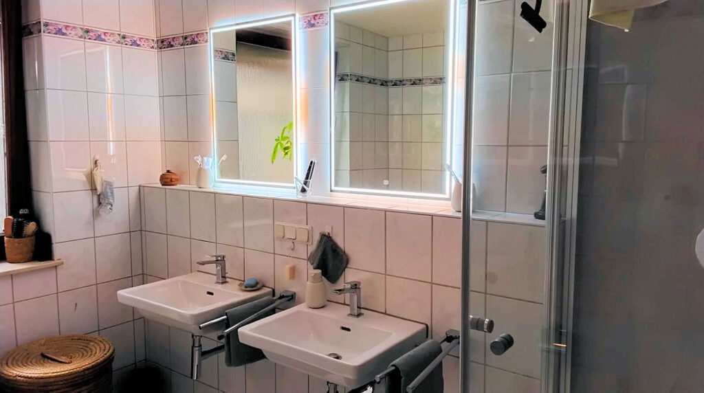 Auf dem Bild ist eine Seite des Badezimmers abgebildet. Zu sehen sind zwei separate weiße Waschtische sowie zwei separate beleuchtete Spiegel über einer Vorwandinstallation. Auf der rechten Seite erkennt man ein Teil der Dusche und auf der linken Seite sieht man ein Teil des Fensterrahmens. Unter dem Fenster befindet sich ein geflochtener runder Korb mit Deckel. Die Wände sind weiß gefliest mit einer Borde im oberen Drittel.
