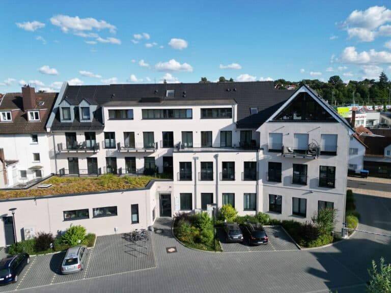 Auf dem Foto erkennt man die Rückseite ein Gebäude welches im Erdgeschoss und 1. Obergeschoss gewerblich genutzt wird. Auf der linken Seite des Gebäudes befindet sich ein eingeschossiger Anbau mit begrüntem Flachdach und mehreren Fenstern. Das zweite Obergeschoss und das Dachgeschoss mit Gauben ist als Wohnraum ausgebaut. Vier Balkone sind im 2. Obergeschoss und zwei Balkone im 1. Obergeschoss zu sehen. Das Gebäude ist symmetrisch mit bodentiefen Fensterflächen auf jeder Etage versehen. Die Fassade ist in zarten Beige-Tönen gestrichen. Im Vordergrund des Gebäudes erkennt man einen gepflasterte Innenhof mit Parkbuchen und Fahrradständern, umrahmt von sehr gepflegten Grünflächen mit Büschen und Sträuchern. Die Zufahrt auf den Innenhof ist durch eine Schranke zur Bahnhofstraße abgegrenzt. Der Himmel ist blau mit zarten weißen Wölkchen.