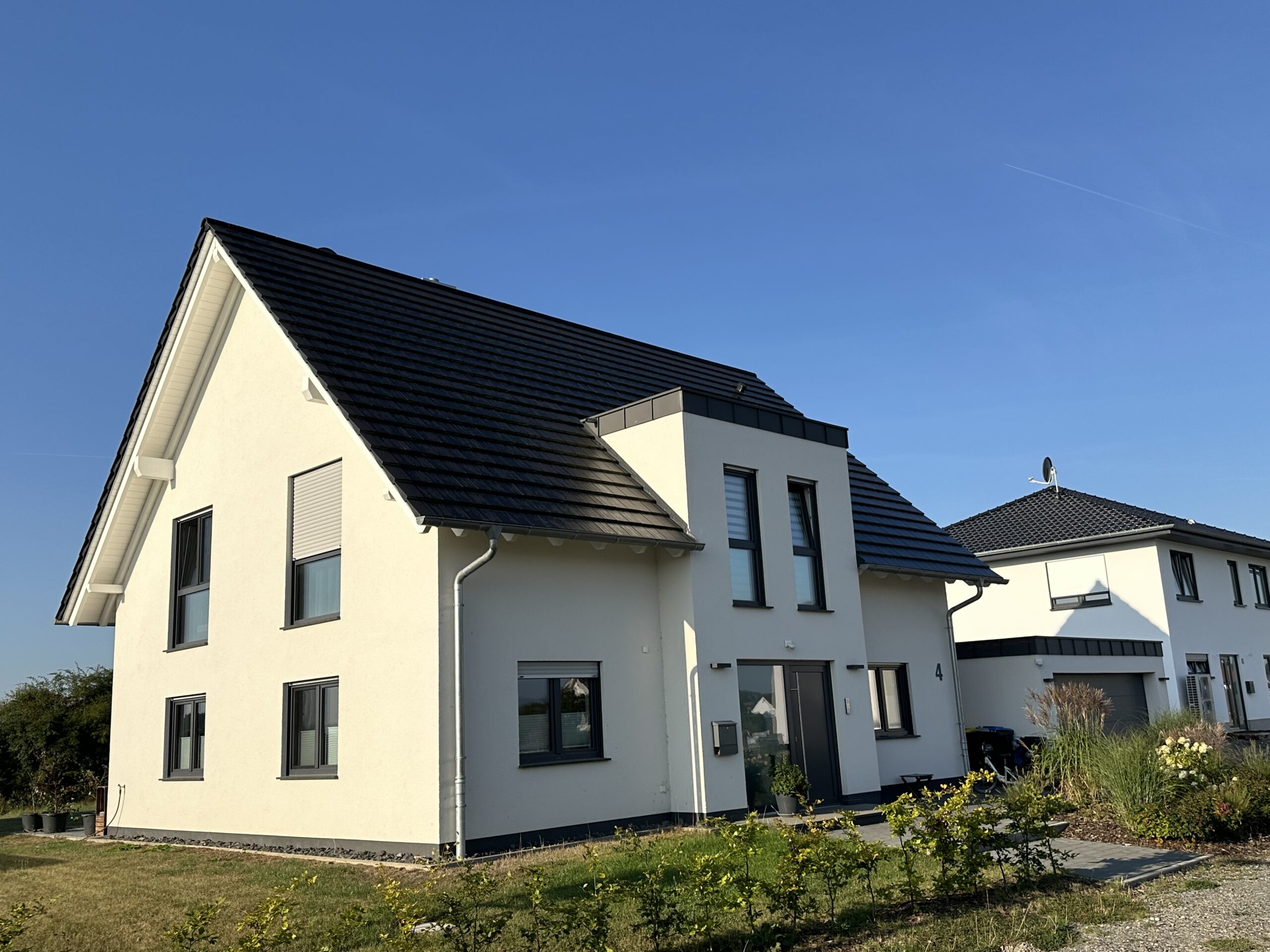 Auf diesem Foto ist ein Neubau eines Einfamilienhauses in zeitgemäßer Architektur mit Erdgeschoss und ausgebautem Dachgeschoss zu sehen. Rechts neben dem Gebäude befindet sich eine Doppelgarage mit Flachdach. Die Fassade von Haus und Garage ist weiß gestrichen, die Fenster, das Garagentor und die Dacheindeckung sind anthrazitfarben. Die Fenster im Obergeschoss der Immobilie sind sämtlich bodentief ausgeführt. Zur Straßenseite befindet sich eine neu gepflanzte Hecke. Der Himmel ist strahlend blau.