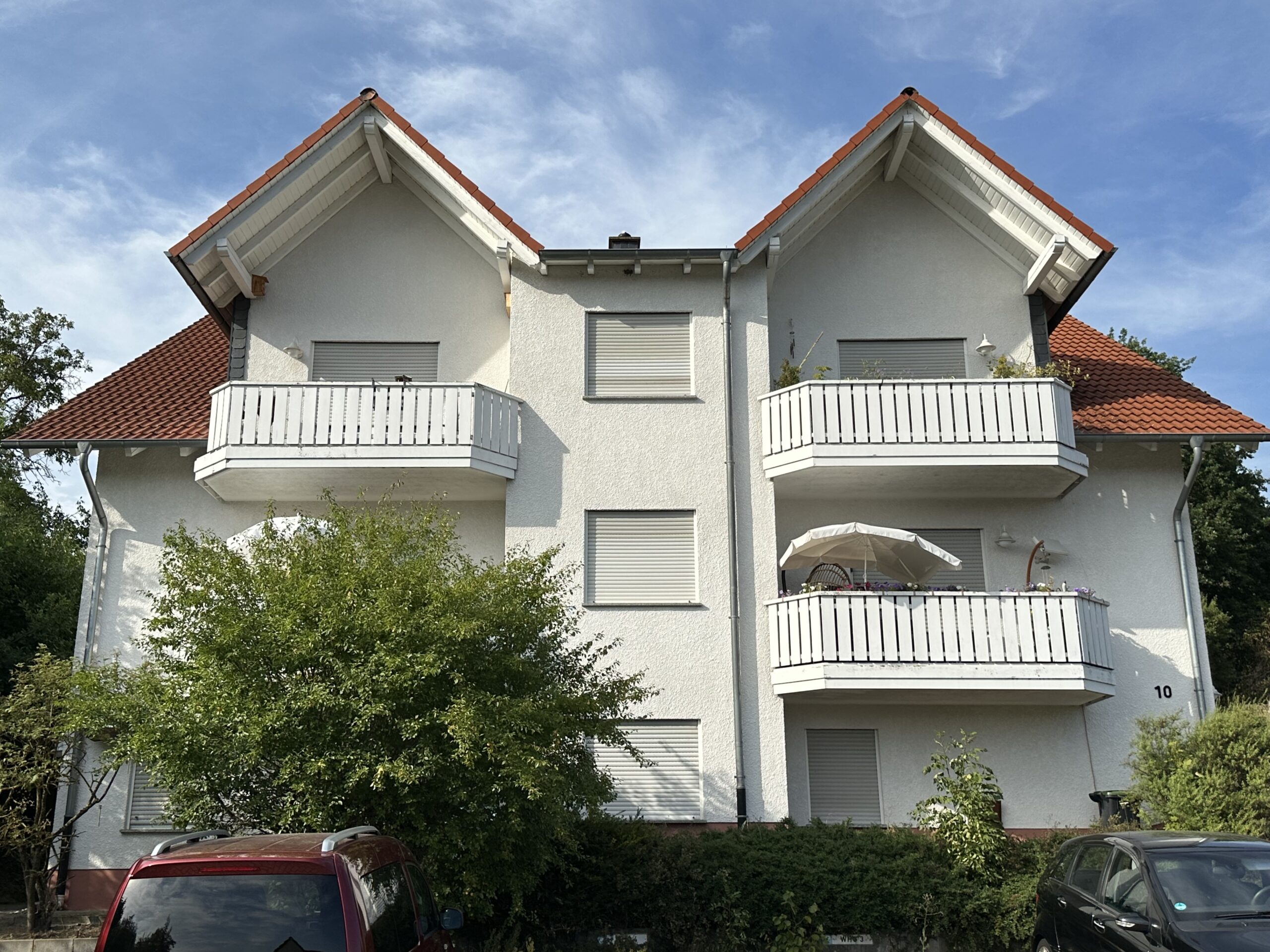 Das Bild zeigt ein dreistöckiges Wohnhaus mit Balkonen und rotem Ziegeldach an einem sonnigen Tag. Im Detail ist ein weißes, modernes Wohnhaus mit drei Stockwerken zu sehen. Das Haus hat ein rotes Ziegeldach und weiße Balkone mit Geländern. Die Fensterrahmen sind ebenfalls weiß. An der Fassade ist die Nummer "10" angebracht. Vor dem Haus befindet sich ein grüner Hang, der durch einen Maschendrahtzaun begrenzt wird.