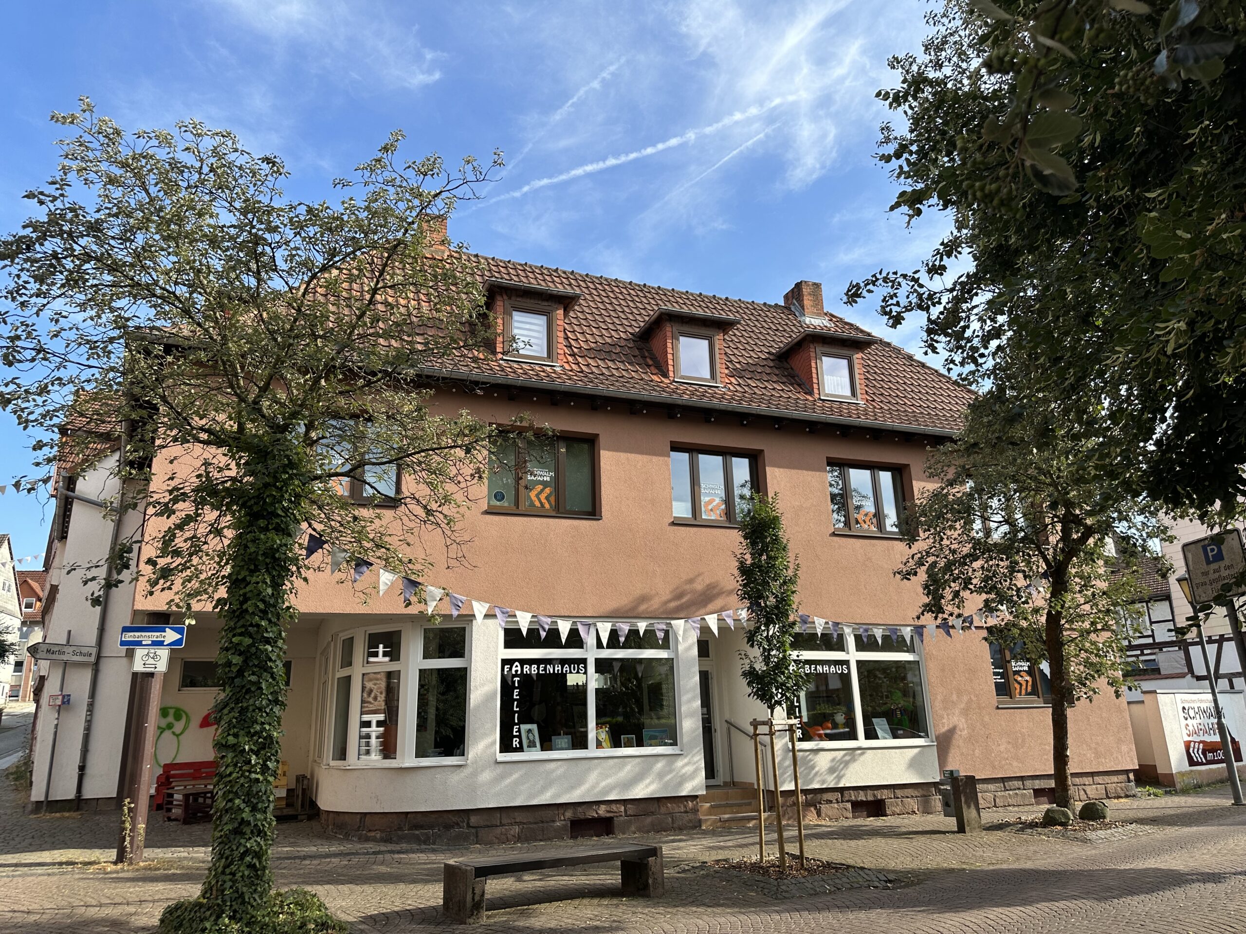 Auf diesem Foto ist ein ein Wohn- und Geschäftshaus zu sehen. Im Erdgeschoss befindet sich ein Atelier, im Obergeschoss eine Fahrschule und im Dachgeschoss eine Wohnung. Die Fassade ist in braun und beige Tönen gestrichen und sieht sehr gepflegt aus. Vor dem Haus befinden sich ein Gehweg mit Sitzbank, sowie eine Einbaustraße mit Parkbuchten. Die Flächen sind sämtlich gepflastert.
